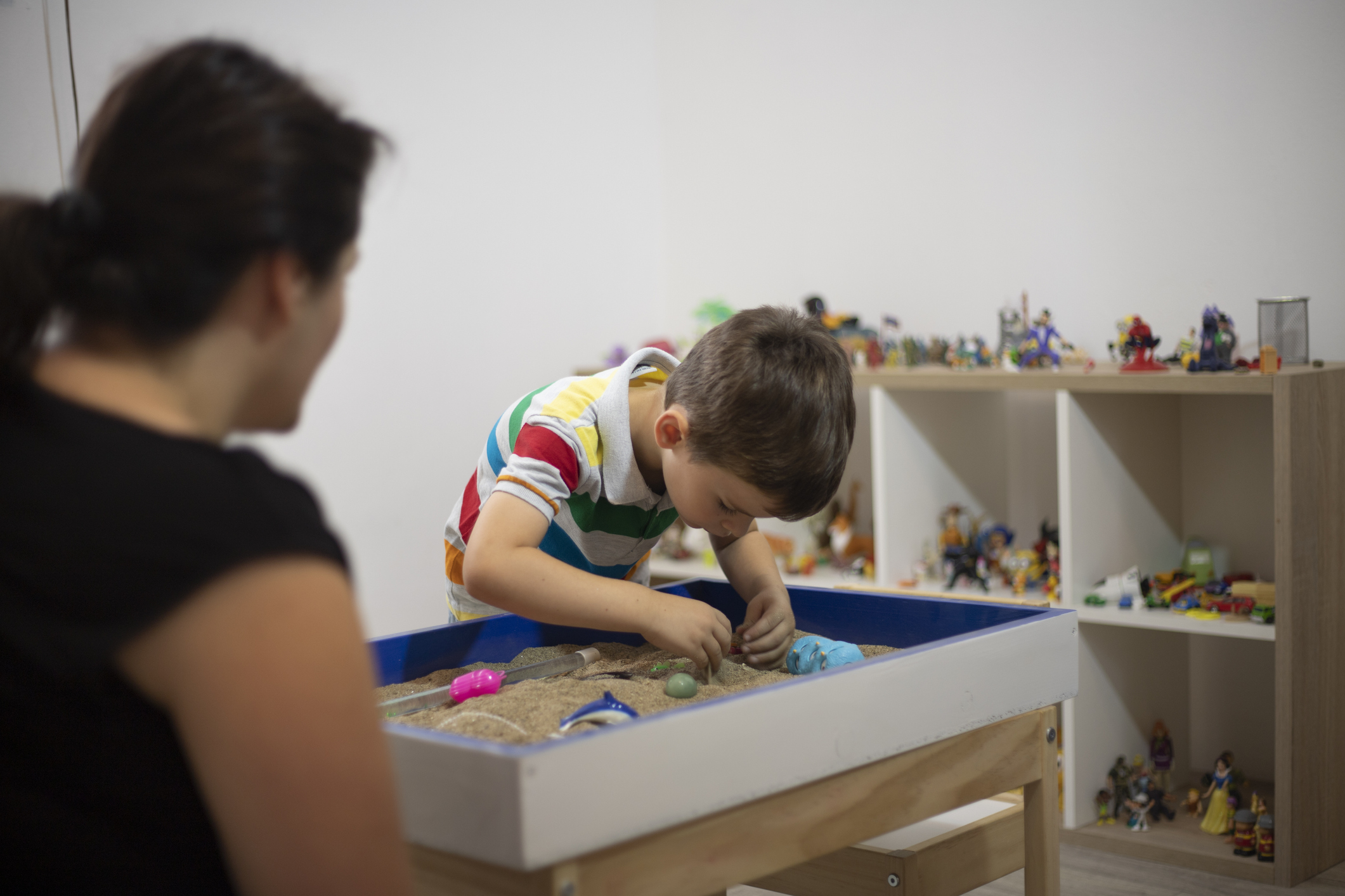 Photo of a woman with her back to the camera. There is a young boy playing in a sand tray with a shelf of toys behind him. 